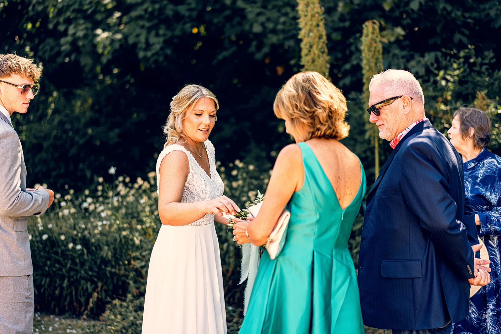 A smiling bride interacting with family members outdoors in a lush garden setting on a bright summer wedding day.