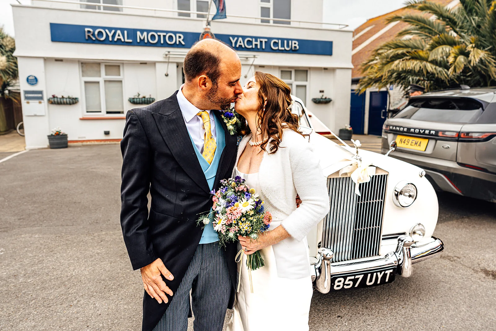 A newlywed couple shares a tender kiss outside the Royal Motor Yacht Club next to a vintage white Rolls Royce wedding car.