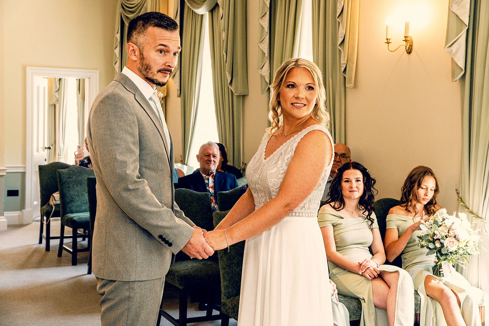 A groom in a grey suit and a bride in a lace-detailed gown hold hands during their indoor civil ceremony while seated guests look on.