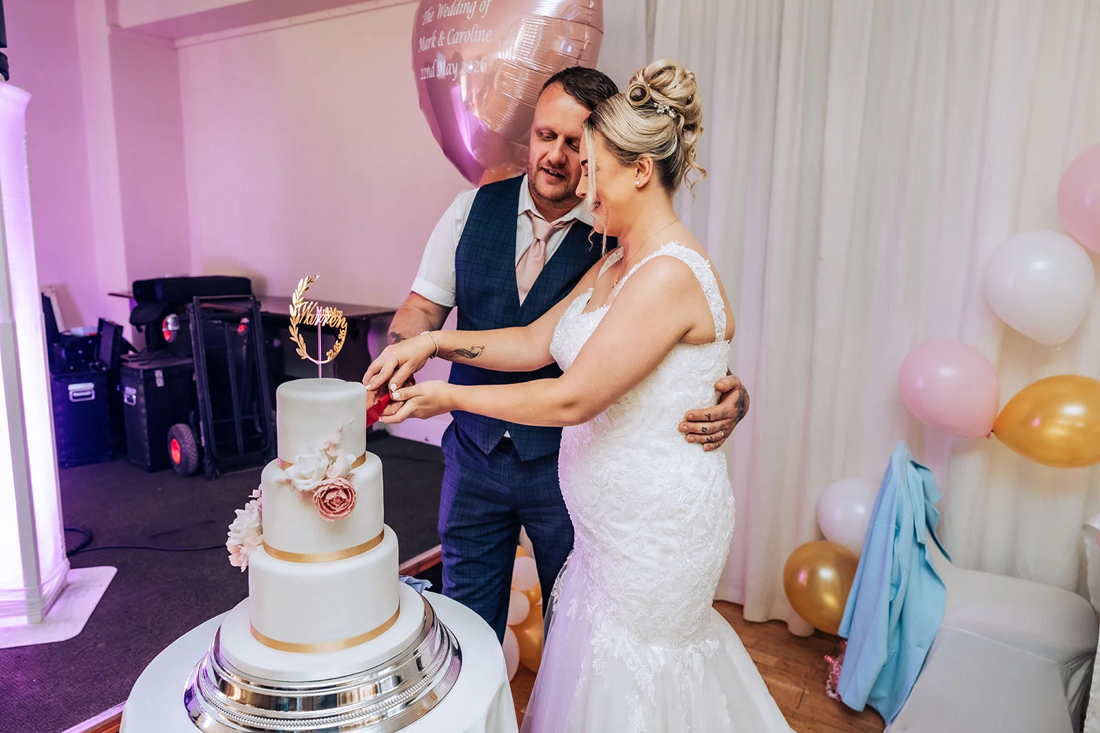 The pregnant bride and groom lovingly hold the knife together to cut their white tiered wedding cake adorned with pink and white floral accents.