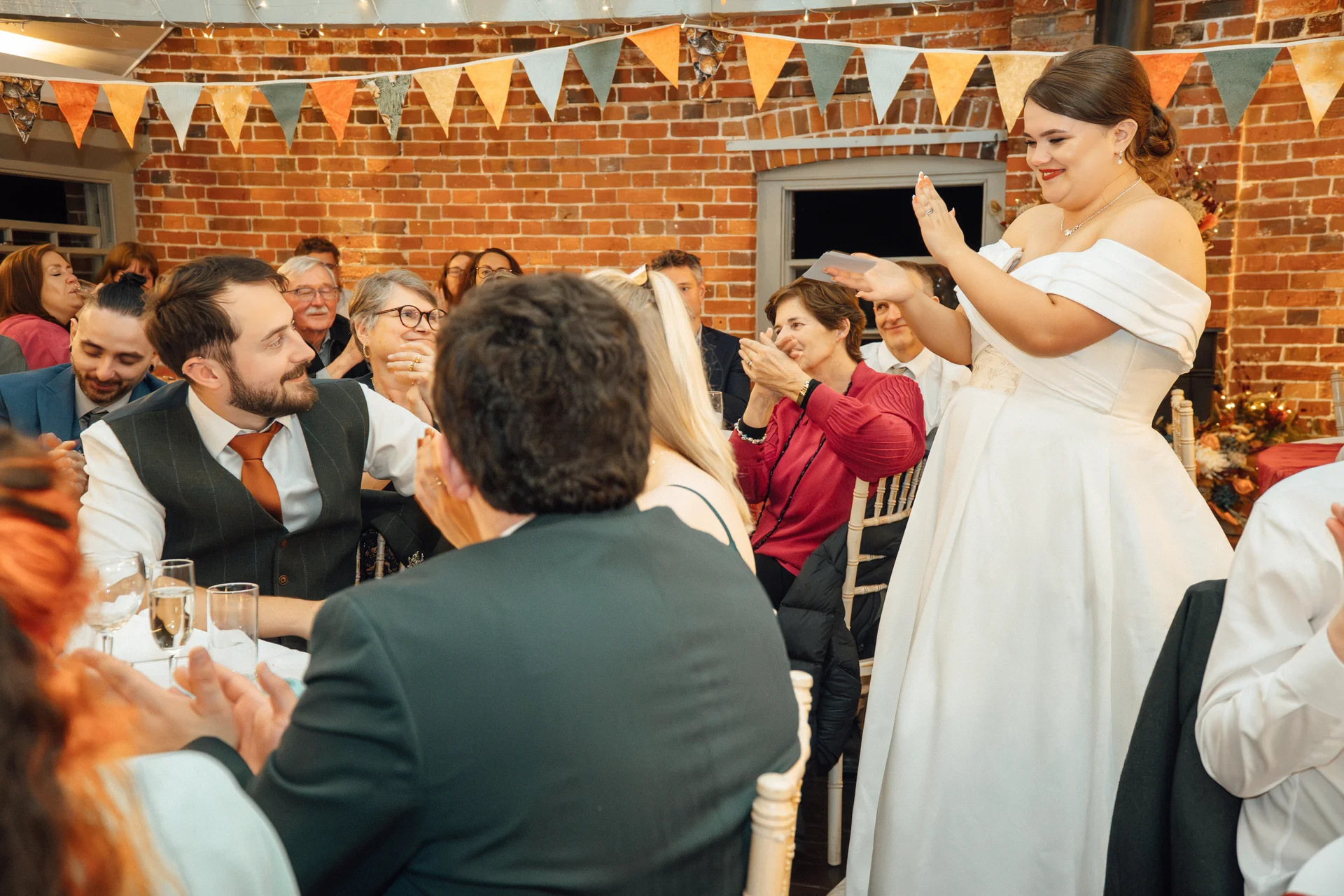 A joyful bride stands to deliver her wedding speech in a rustic brick venue decorated with colourful bunting while seated guests applaud and smile.