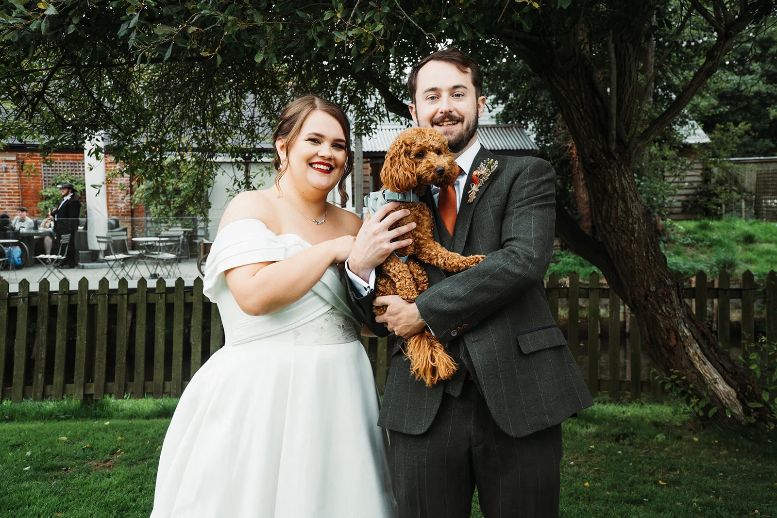 A smiling bride and groom pose outdoors in a garden setting, with the groom proudly holding their fluffy brown dog.