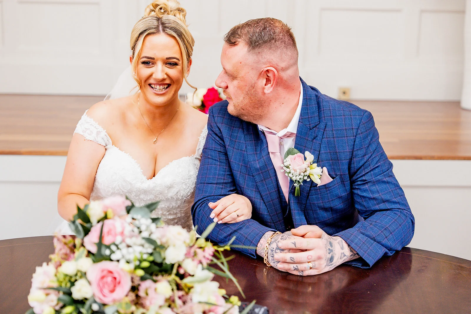A newly married couple laughing together as they sit at a wooden table during their wedding ceremony.