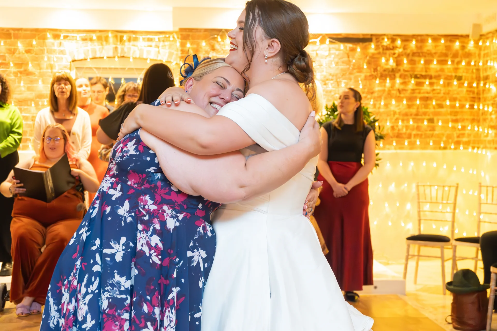 Two brides joyfully embrace each other tightly in an indoor venue adorned with warm fairy lights and exposed brick walls.