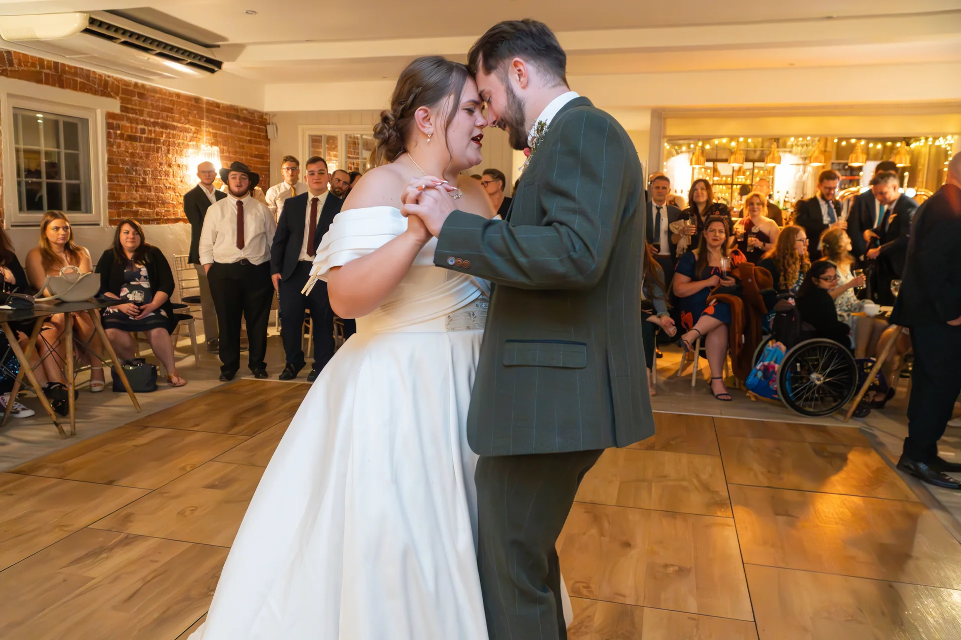 Newlyweds share an intimate nose-to-nose moment during their first dance on a wooden dance floor, surrounded by watching guests in a venue with exposed brick walls.