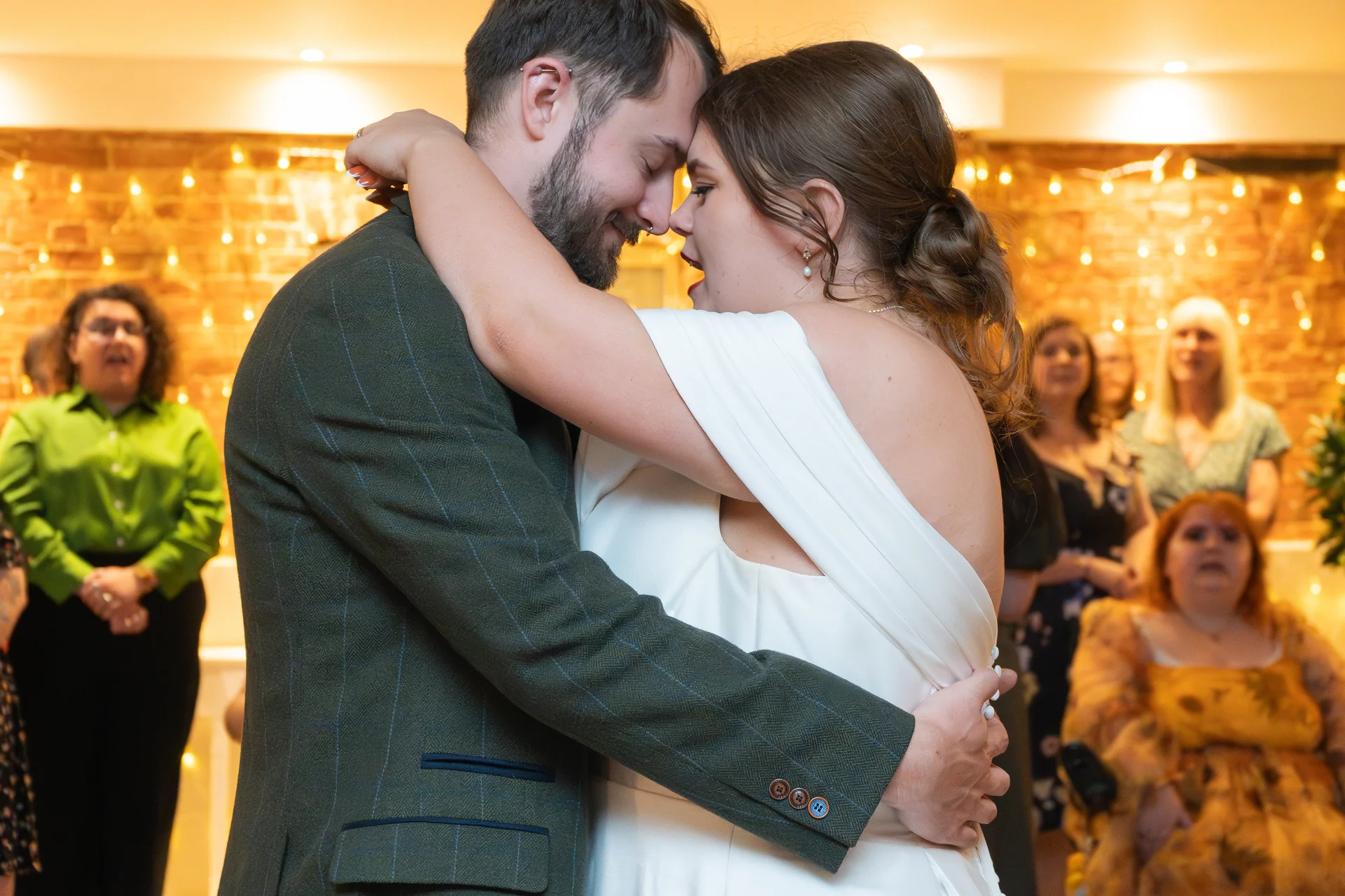 A bride and groom share an intimate embrace and touch noses during their first dance while wedding guests watch in the background.