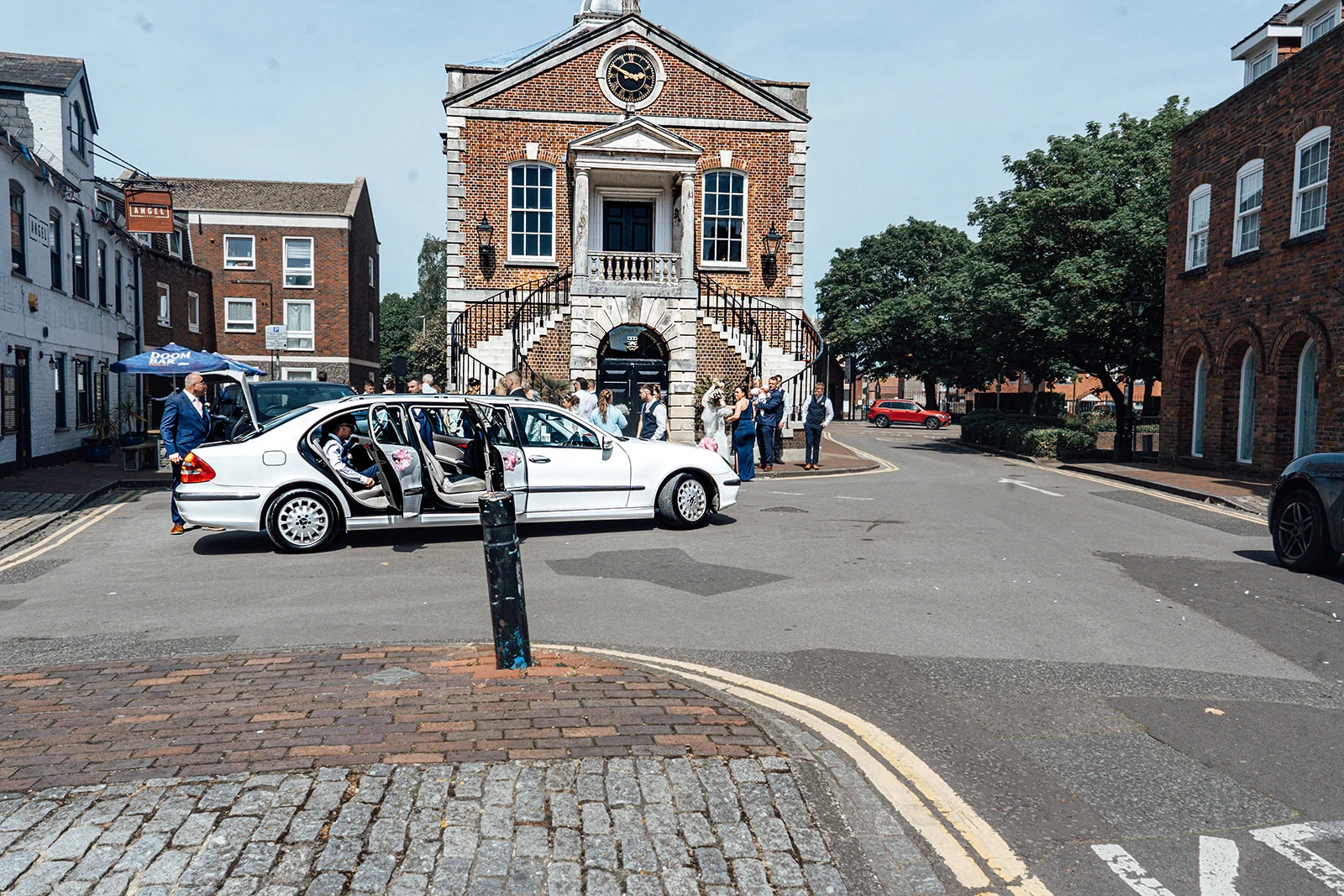 A white wedding car arrives outside a historic brick town hall on a bright sunny day as guests gather for the ceremony.