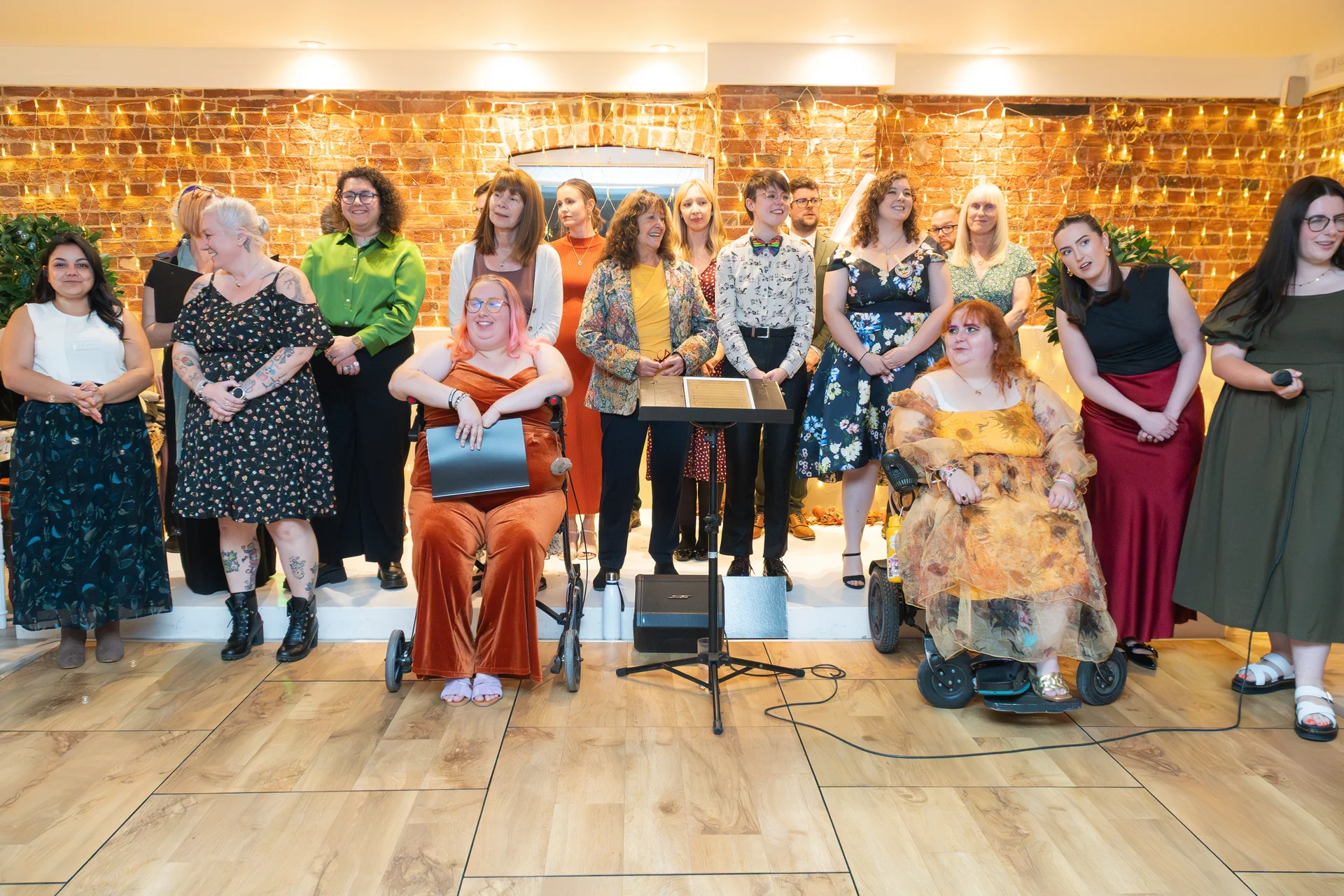 A diverse group of wedding guests and performers stand and sit smiling in front of an exposed brick wall decorated with warm fairy lights.