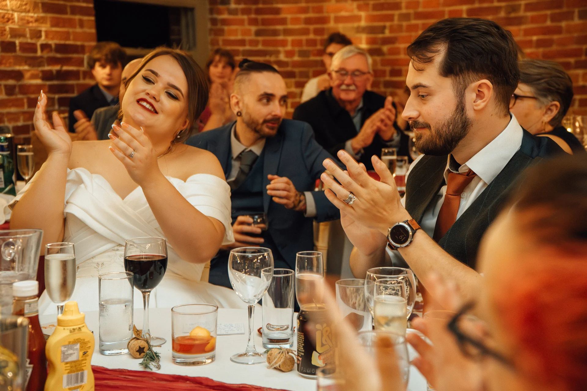 The bride and groom smile and clap happily while seated at a dining table during wedding speeches in a rustic brick venue.