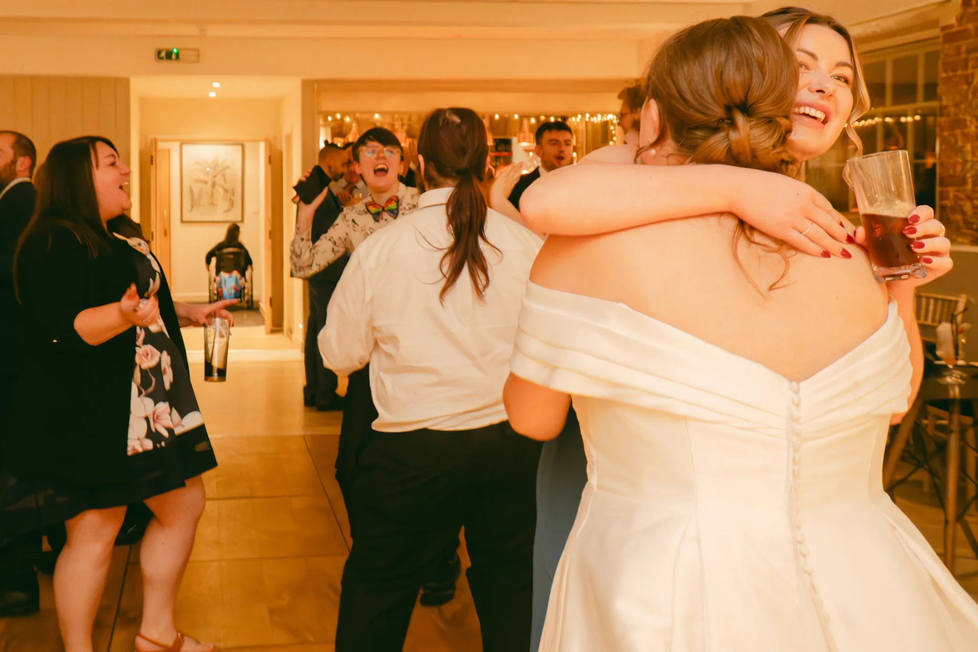 Two brides in an enthusiastic embrace share a joyous laugh on the dance floor surrounded by celebrating wedding guests holding drinks.