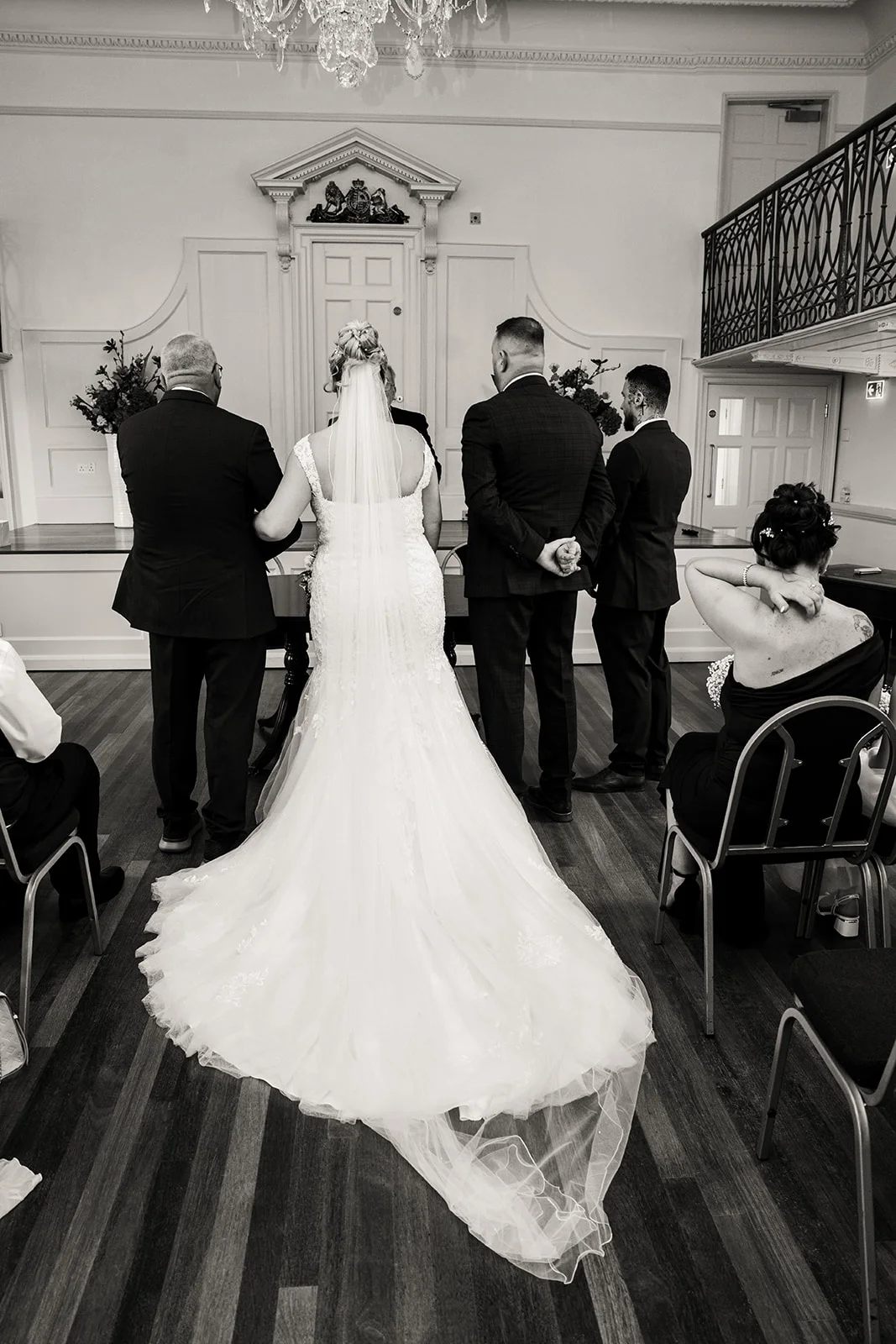 A back view of a bride in a lace a-line dress and chapel veil standing at the registry office table with her partner and guests during the marriage ceremony.