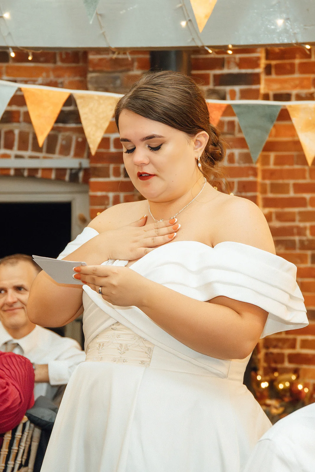 A tearful bride delivers her wedding speech while holding a note to her chest against an exposed brick wall adorned with bunting.