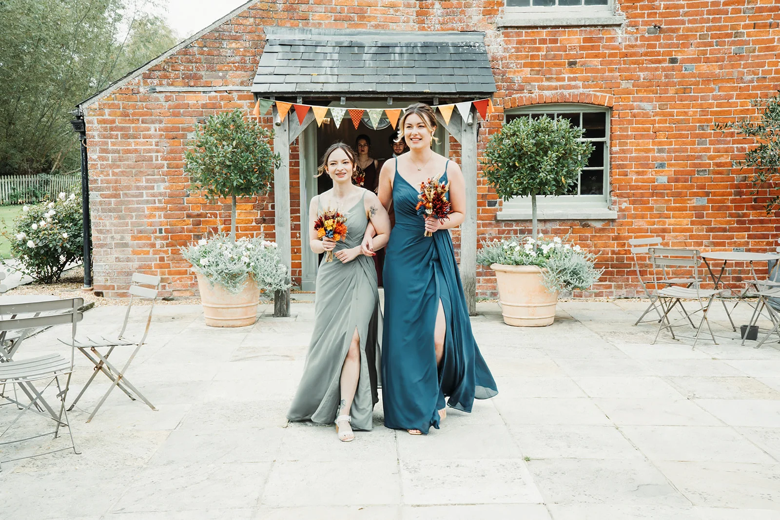 Two bridesmaids in sage and teal dresses walk arm-in-arm out of a brick building featuring colourful bunting and potted trees.