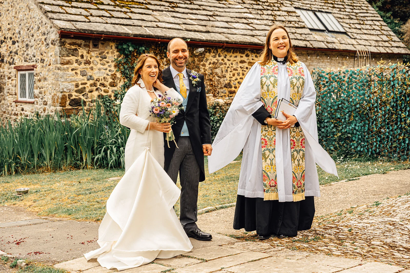 A smiling bride holding a bouquet and groom standing next to an officiant in clerical vestments outside a rustic stone building.
