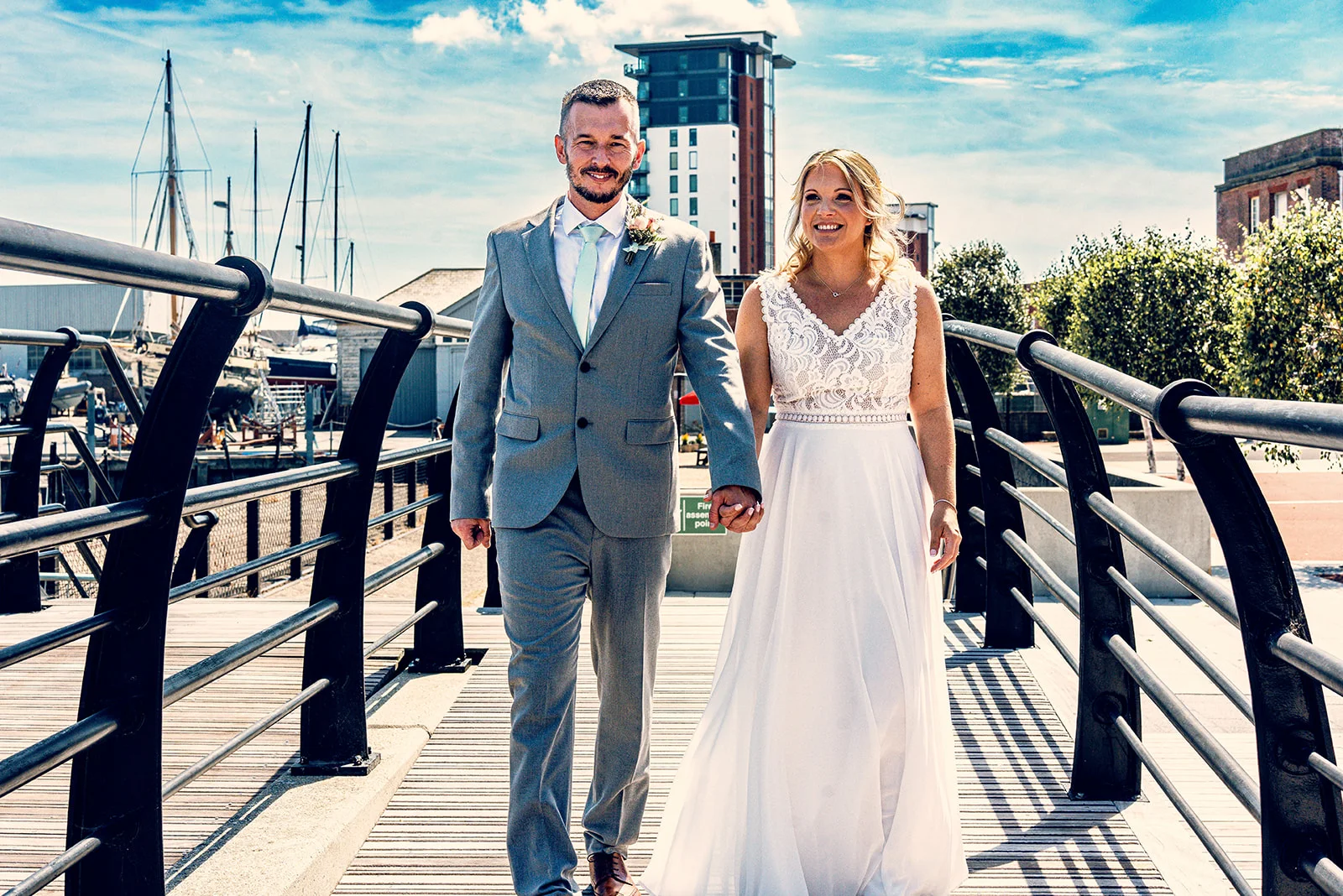 A newlywed couple holds hands while walking together across a modern footbridge near a scenic harbour on a sunny day.