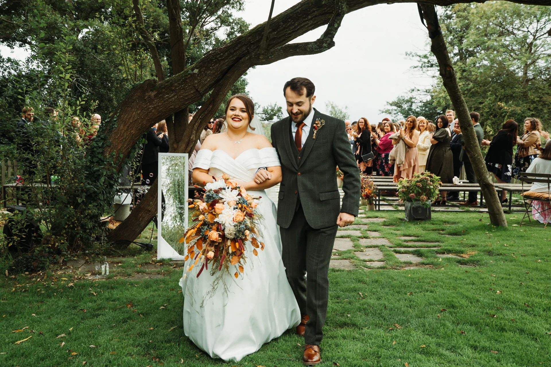 A smiling newlywed couple walks hand in hand down the aisle through a lush garden setting following their outdoor ceremony.