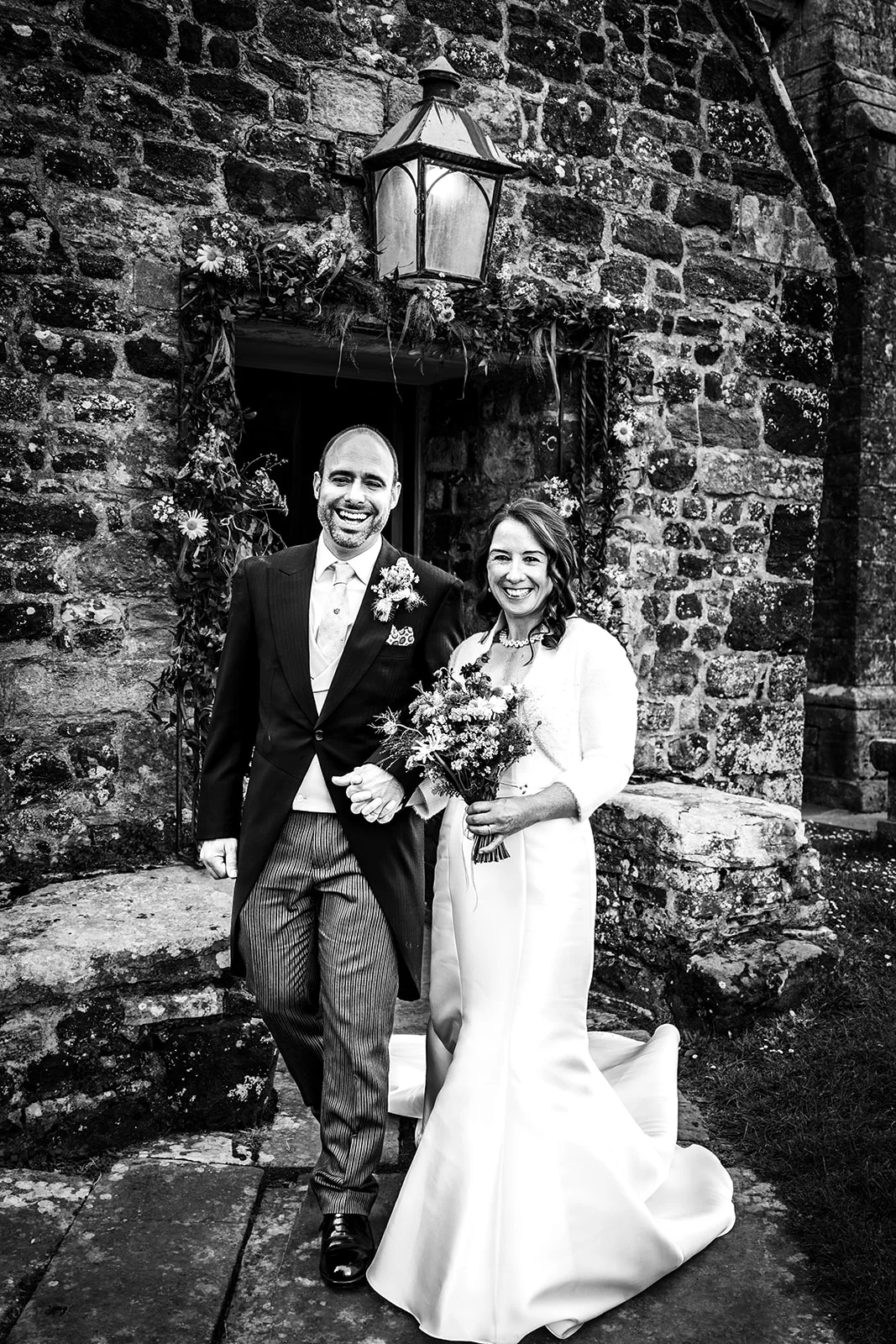 A joyful newlywed couple walks hand in hand out of a stone church doorway, with the groom wearing a morning suit and the bride holding a bouquet.
