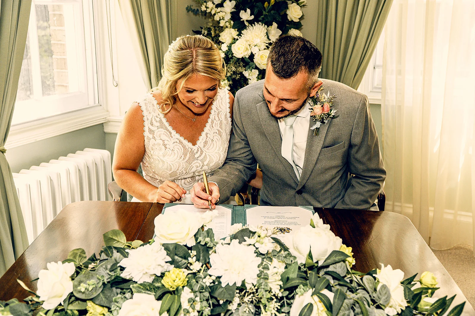 A smiling bride and groom sit together at a wooden table, signing the marriage certificate amidst an arrangement of white roses and green foliage.