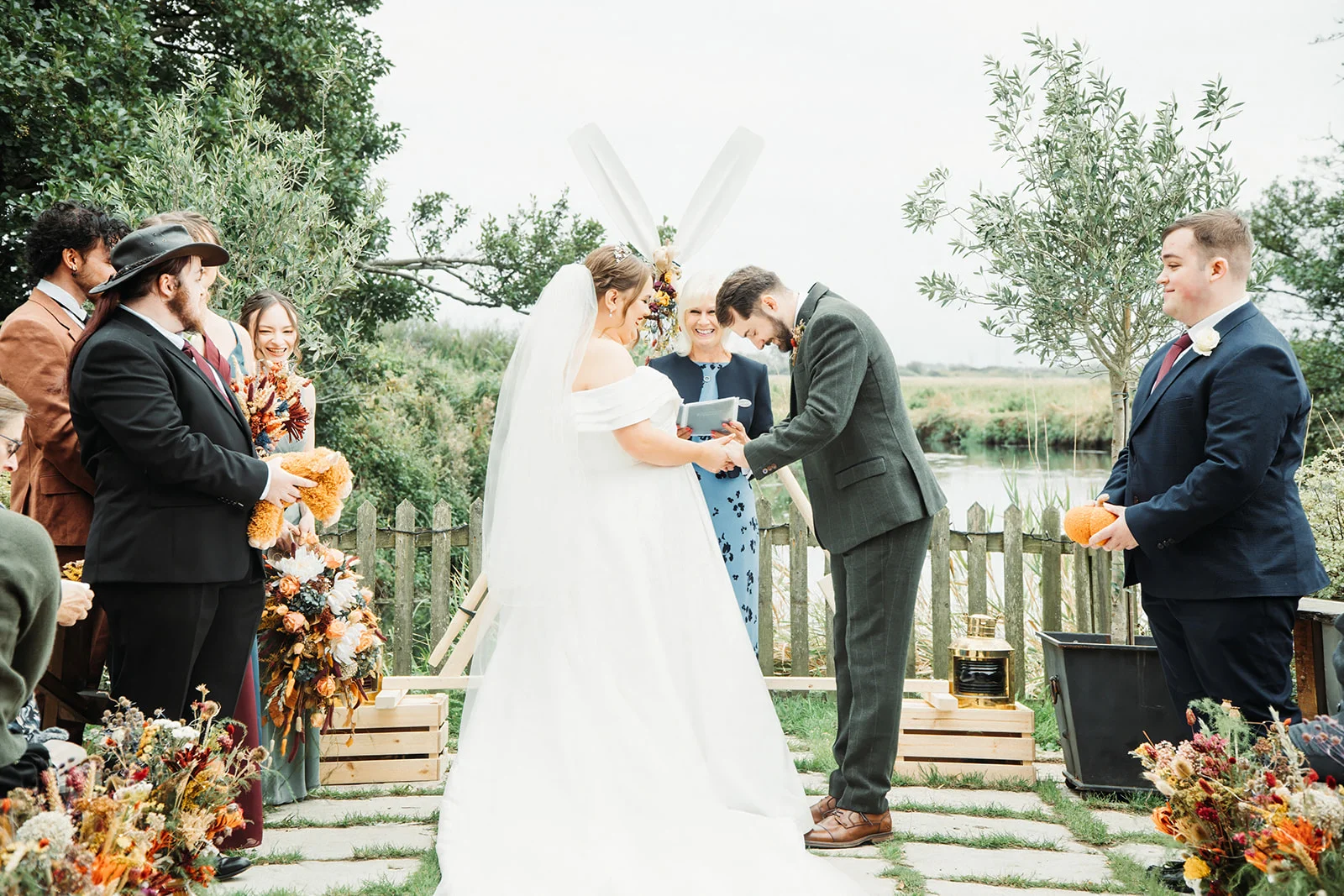 The bride and groom exchange rings during an outdoor lakeside ceremony, officiated by a celebrant and flanked by their wedding party.