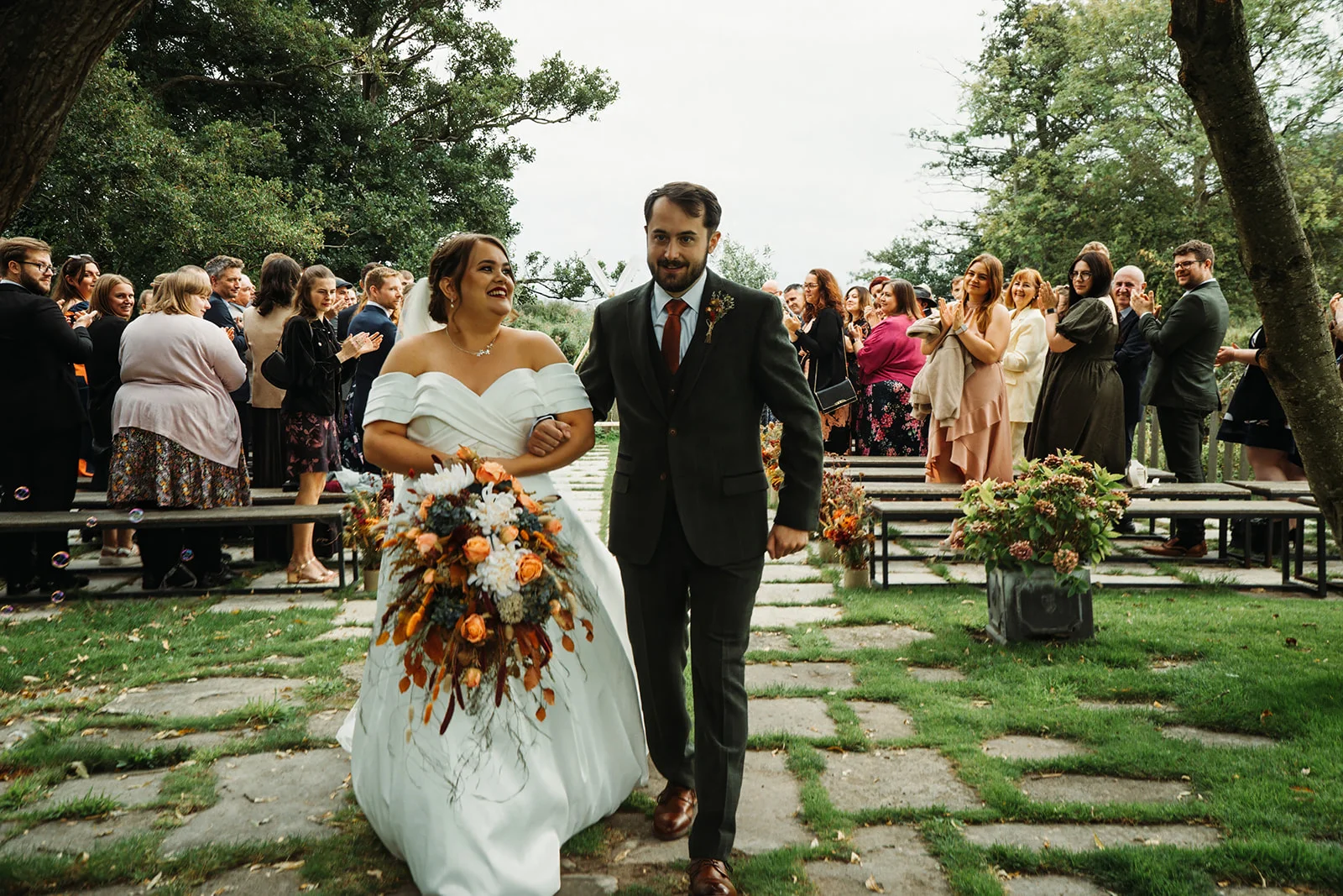 Newlywed bride and groom joyfully walk back down the stone-paved aisle together after their outdoor wedding ceremony, surrounded by cheering guests.