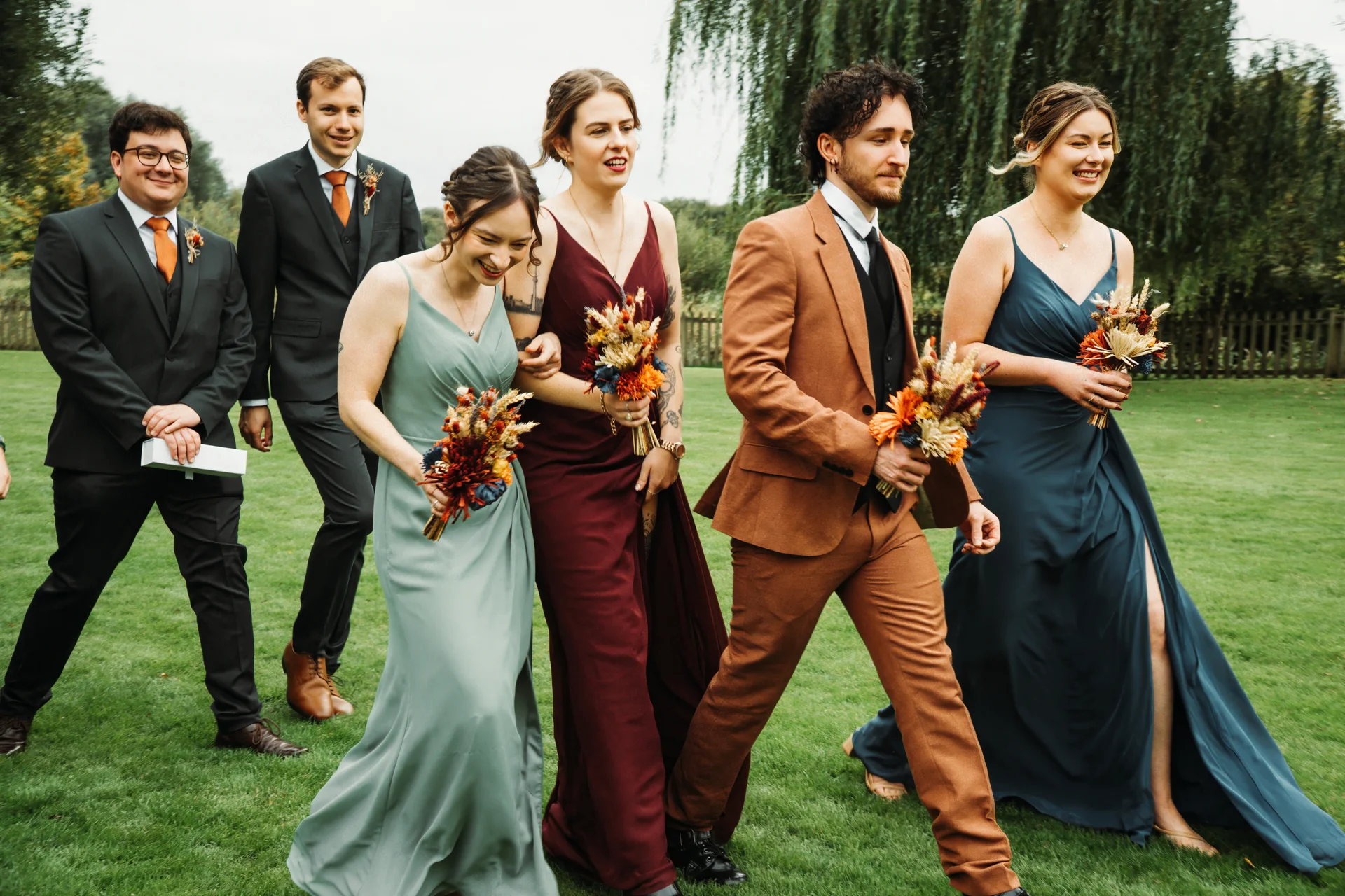A joyful wedding party of bridesmaids in mismatched sage, burgundy and teal gowns alongside groomsmen in suits walk across a lush green lawn holding dried flower bouquets.