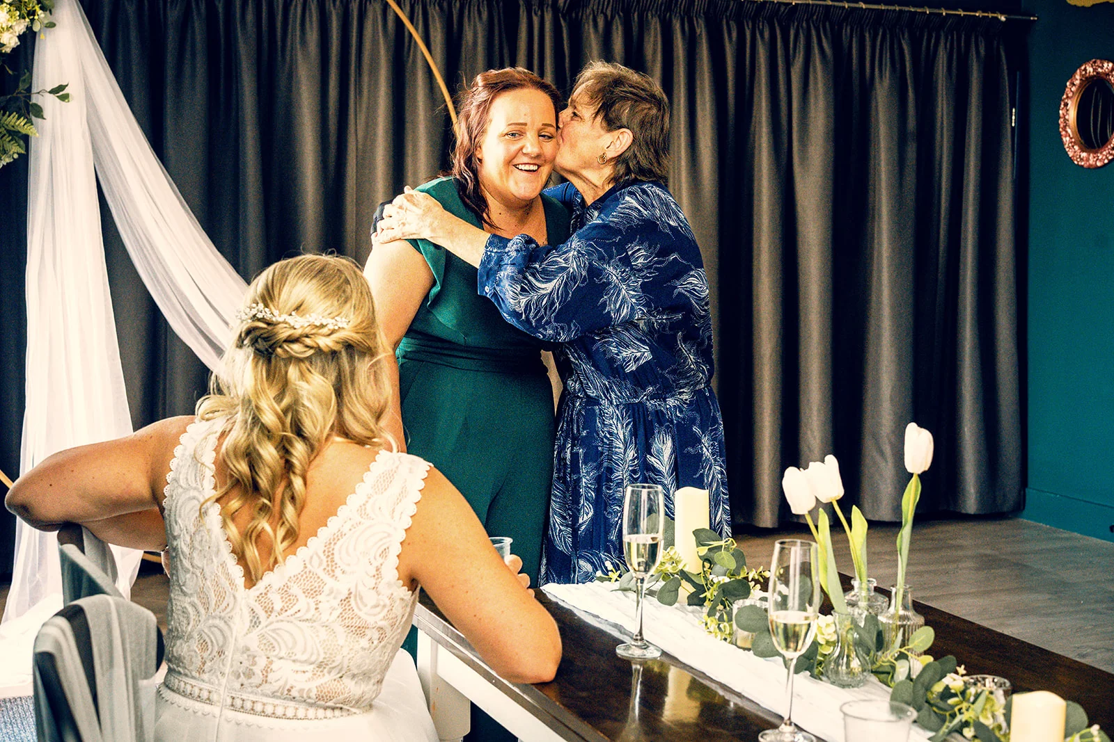 An older guest joyfully kisses a smiling woman while a bride in a lace dress looks on from a decorated wedding breakfast table.