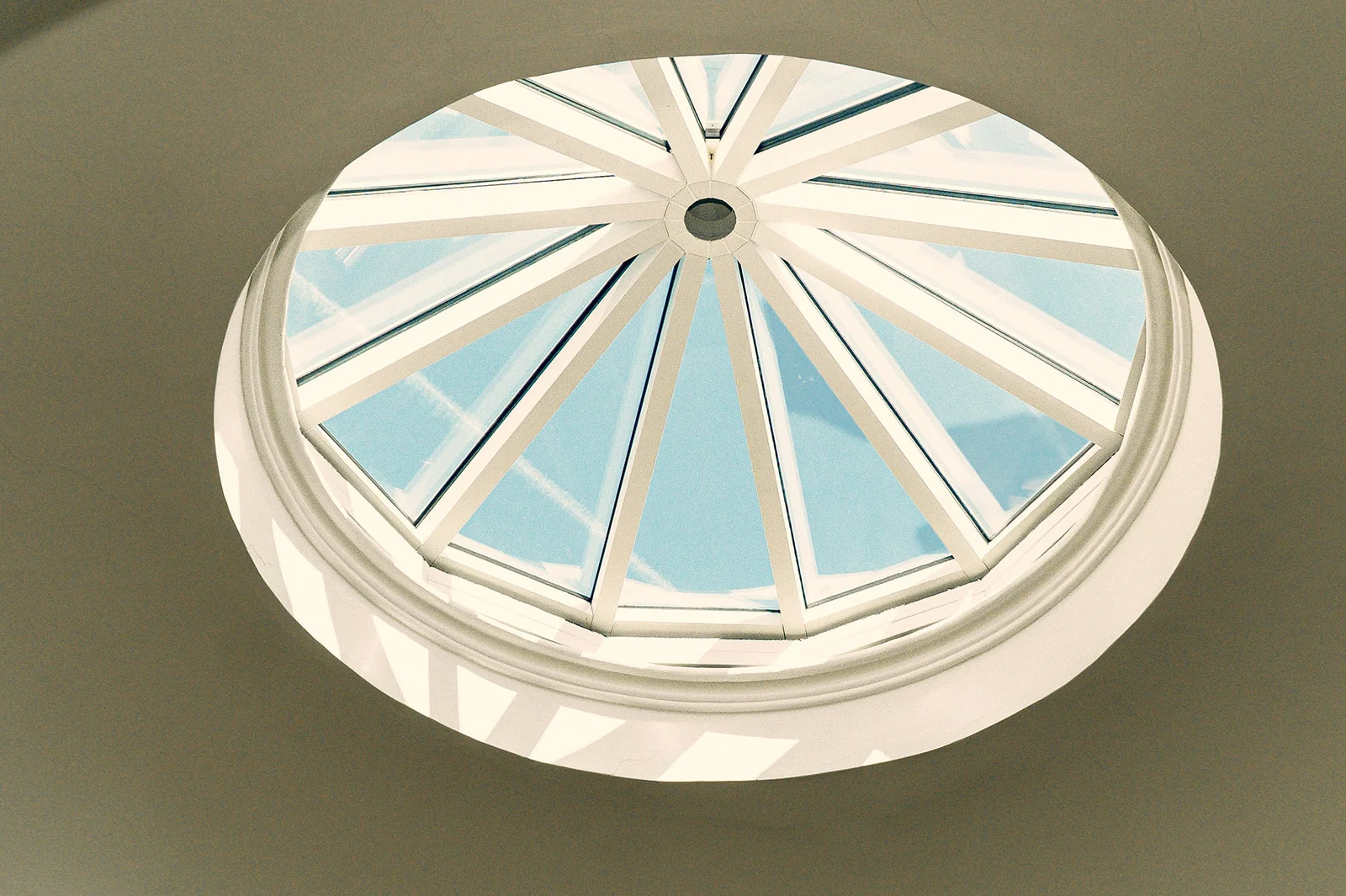 A low angle, symmetrical shot captures a large circular glass skylight set into a pale ceiling, revealing a bright blue sky above.