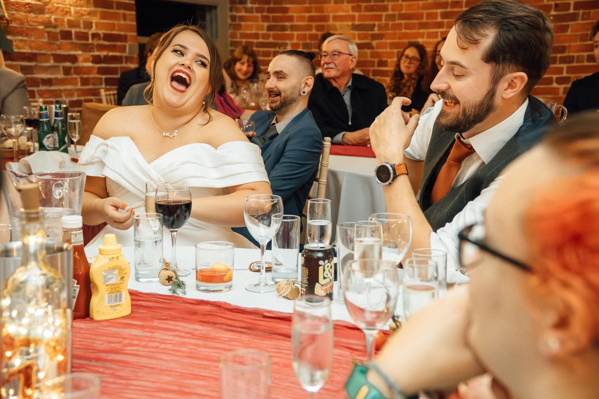 A joyful bride laughs heartily at the wedding breakfast while seated next to the groom in a rustic venue with exposed brick walls.