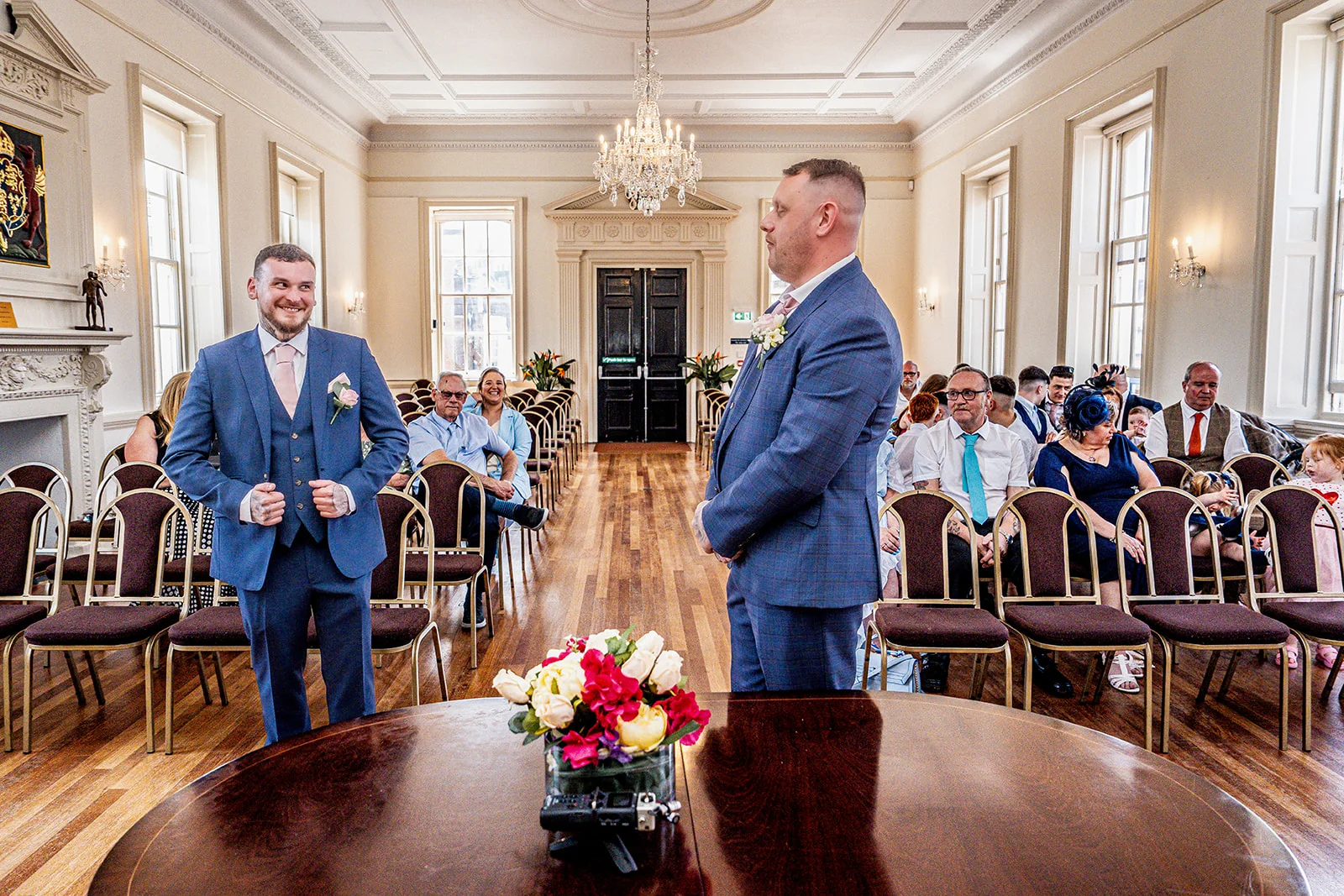 Two grooms in matching blue suits smile happily during their civil ceremony inside a grand, wood-floored hall filled seated guests.