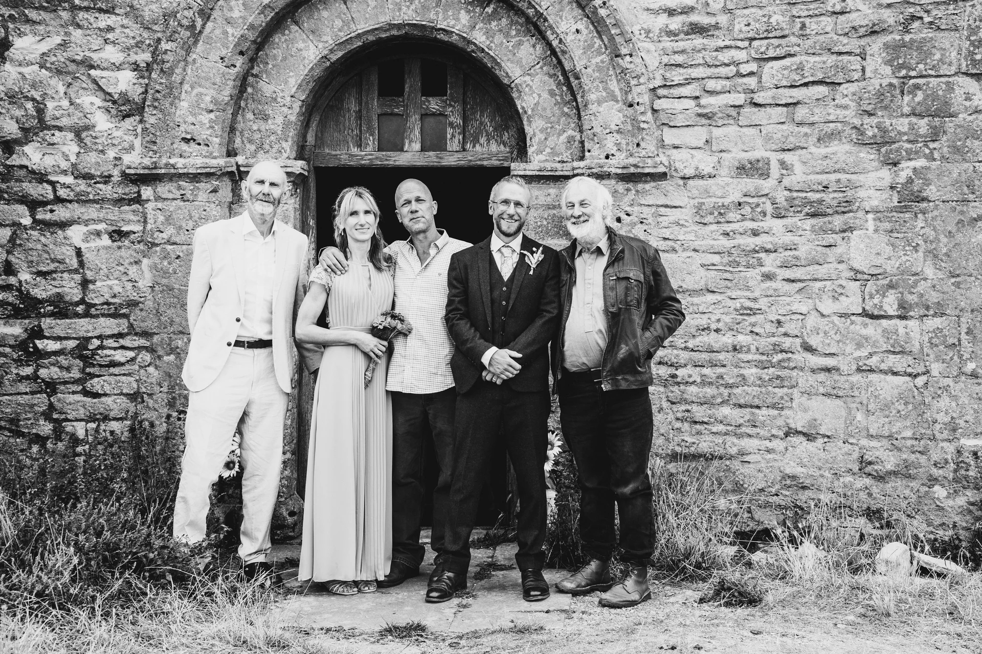 A black and white group portrait of five adults posing together outside a rustic stone building with an arched wooden doorway.