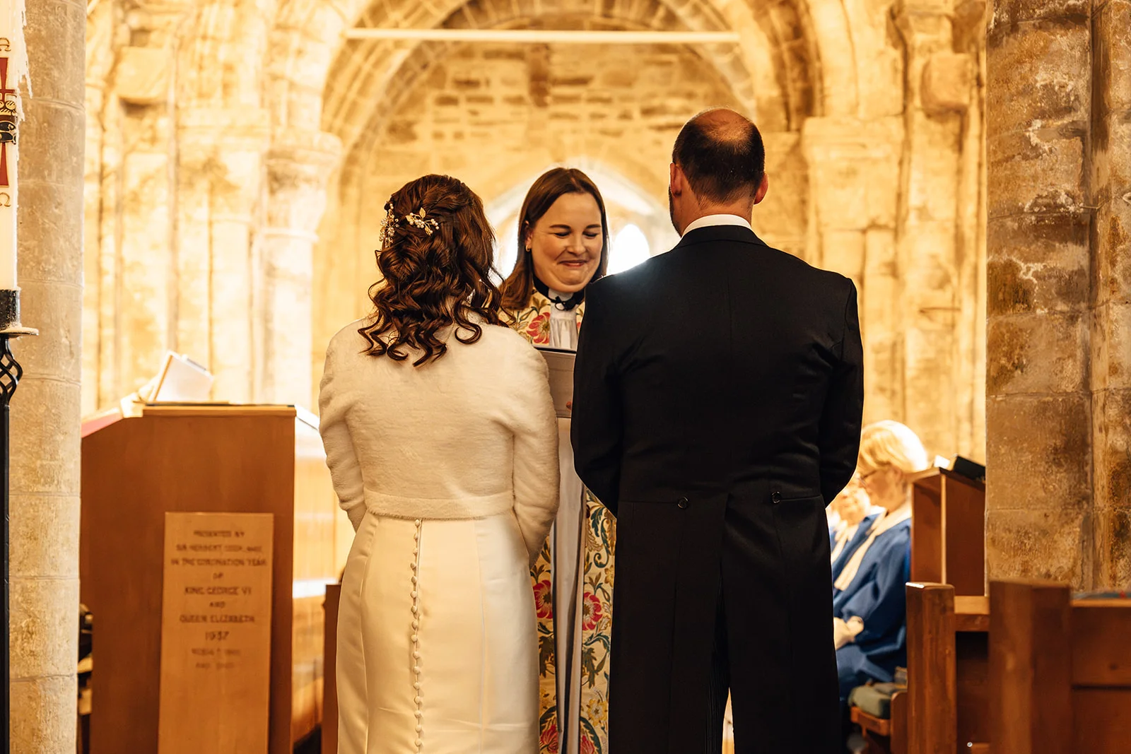 A bride and groom stand at the altar during a church wedding ceremony, viewed from behind as they face the celebrant beneath an ancient stone archway.