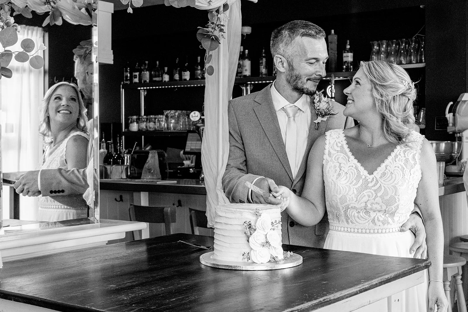 A smiling bride and groom cut their white tiered wedding cake together inside a pub, with their reflection captured in a nearby mirror.