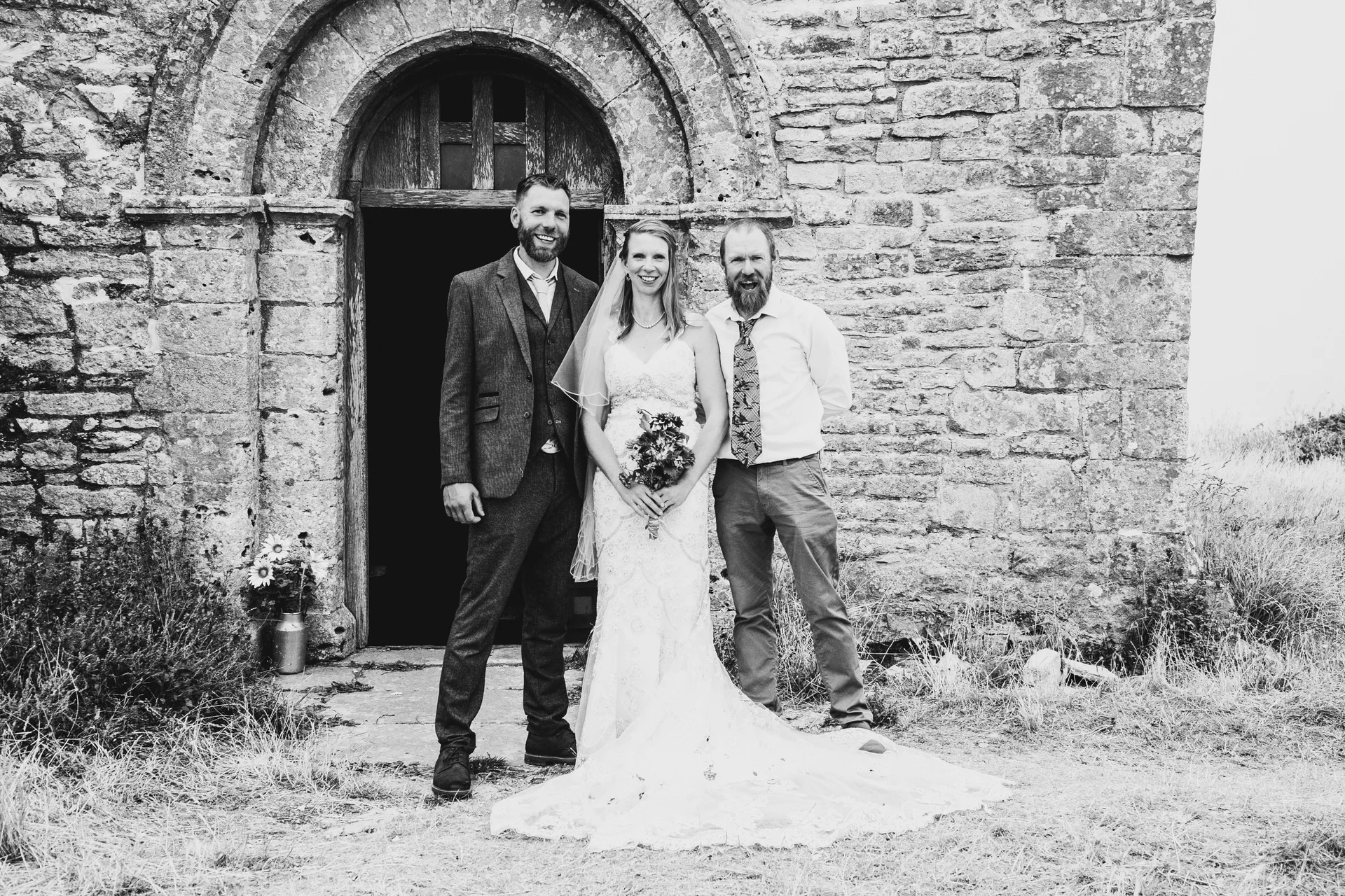 A bride in a lace wedding gown holding a bouquet stands outside a rustic stone chapel door flanked by two smiling men in suits.