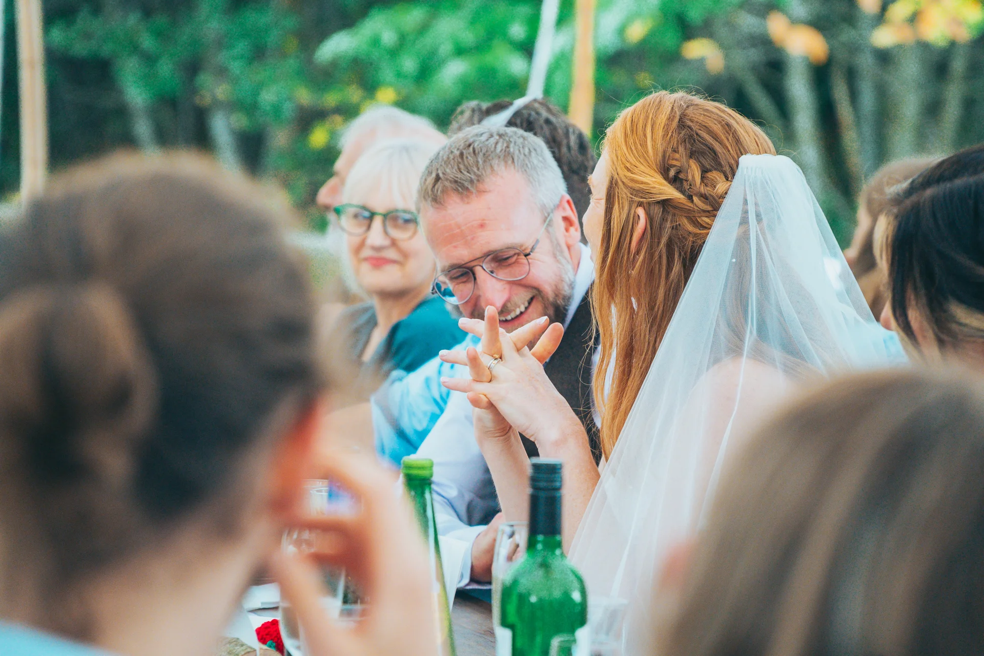 A smiling bride with red hair and a veil shares a joyful moment with the groom at the wedding breakfast while holding hands across the table.