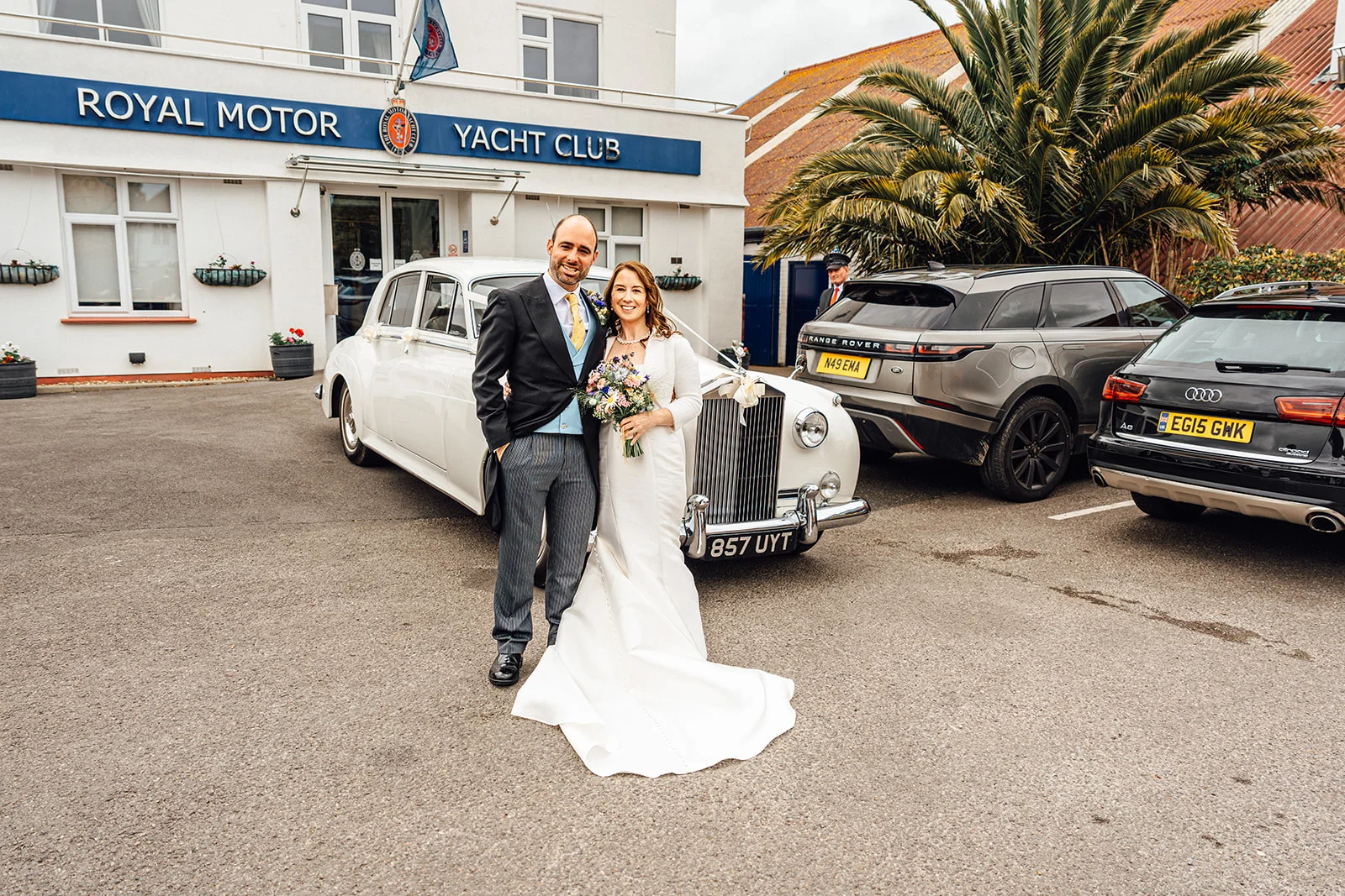 A newlywed couple poses proudly in front of a white vintage wedding car outside the Royal Motor Yacht Club, with the bride holding a delicate floral bouquet.