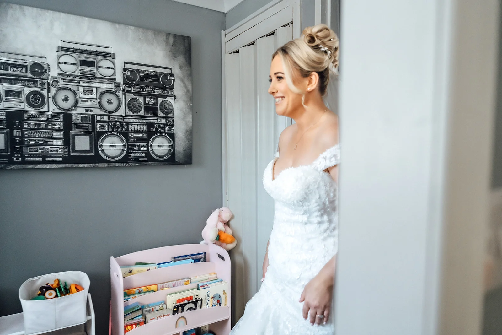 A smiling bride wearing a textured off-the-shoulder wedding gown and an updo hairstyle stands indoors during morning preparations.