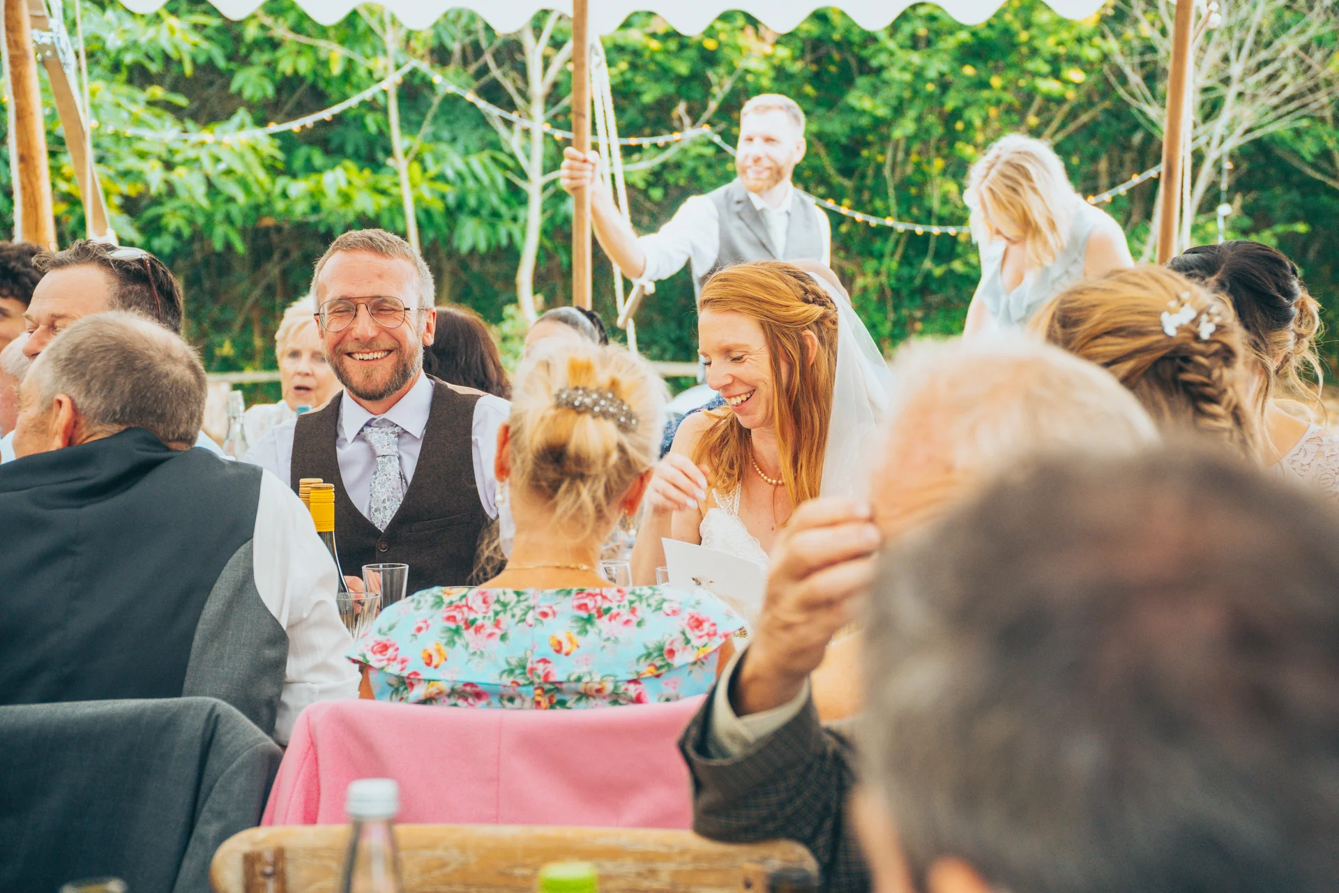 A smiling groom and bride enjoy the wedding breakfast inside a marquee surrounded by laughing guests during speeches.