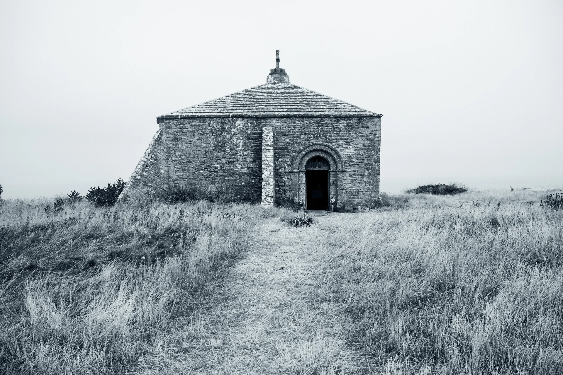 An ancient stone chapel stands isolated amidst overgrown grass under a moody, overcast sky, captured in a striking black and white photograph.