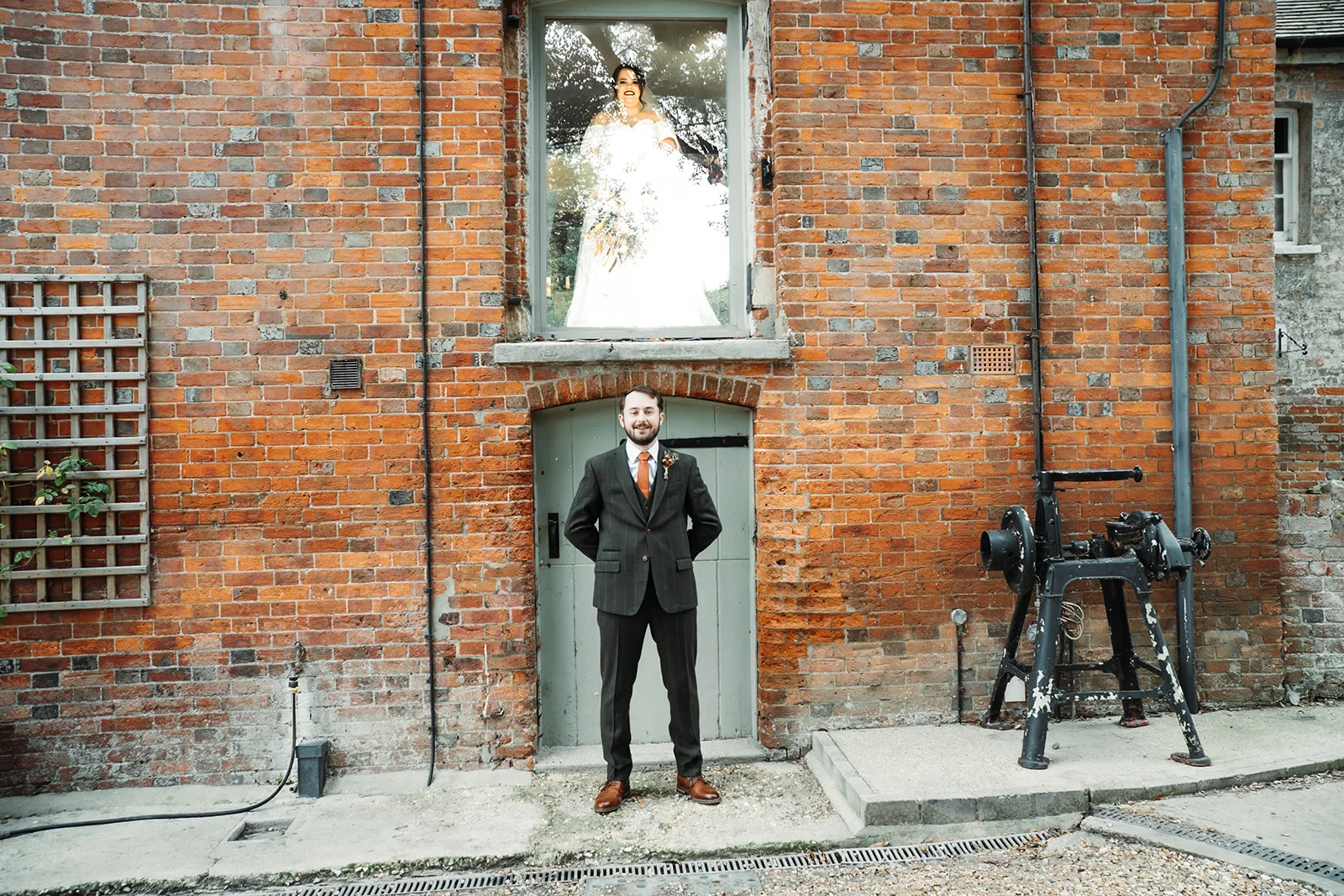 A groom poses outside against a rustic red brick wall while the bride looks down from an upper window.