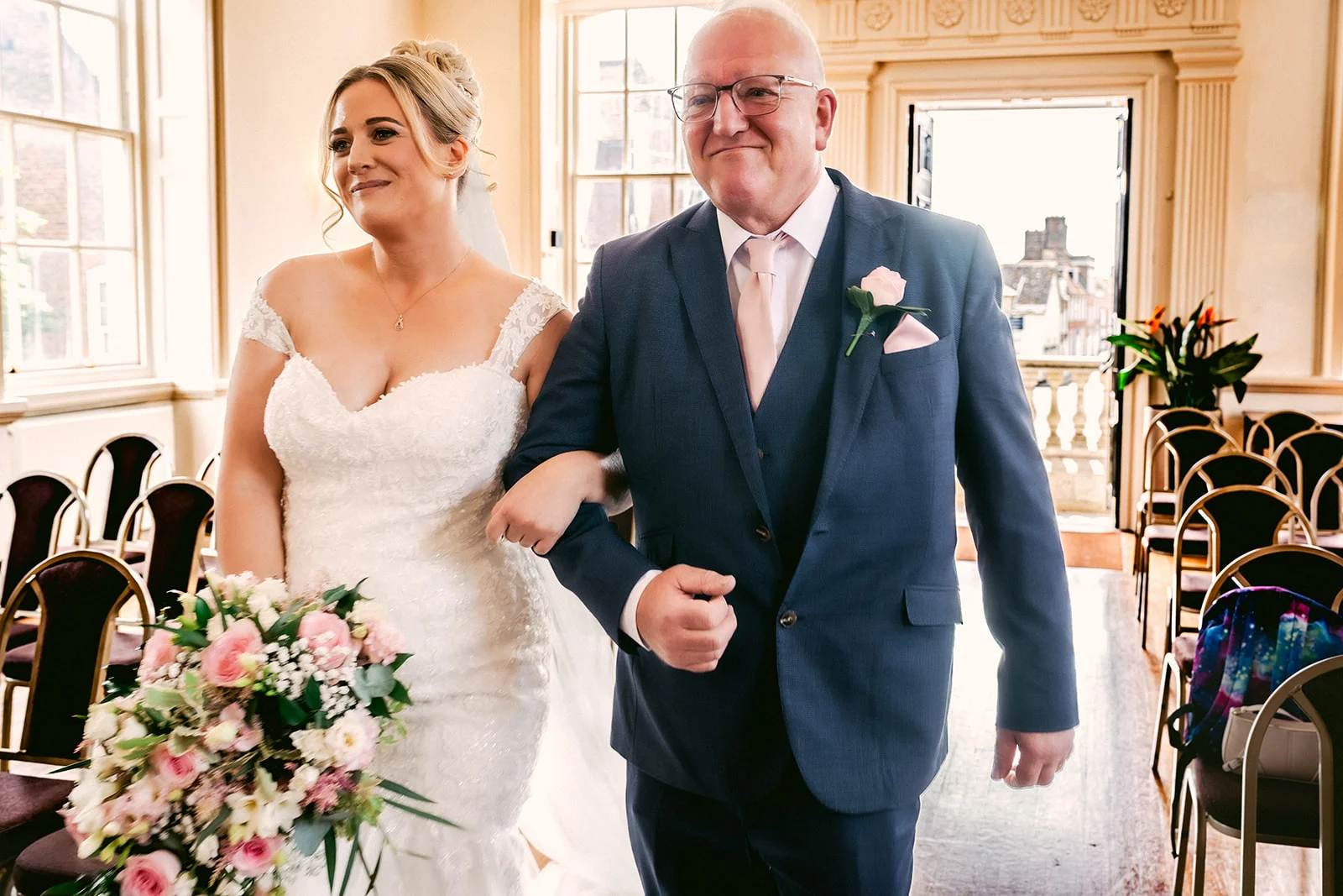 A smiling bride holding a pink and white floral bouquet walks arm in arm with her father in a blue suit inside a light-filled ceremony venue.