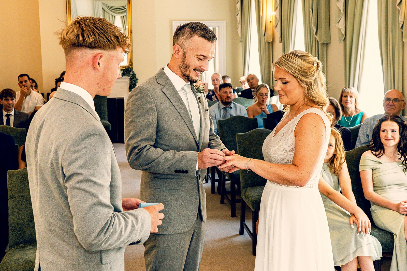 The groom places a ring on the bride's finger during a civil ceremony while guests watch attentively from seated rows.