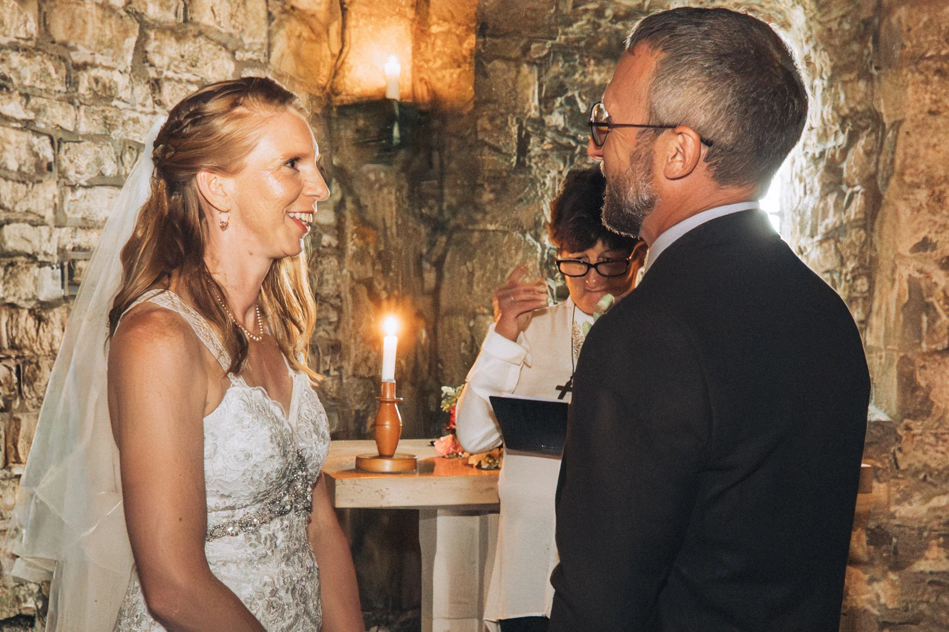 A smiling bride and groom exchange vows in an intimate ceremony inside a historic stone venue illuminated by candlelight.