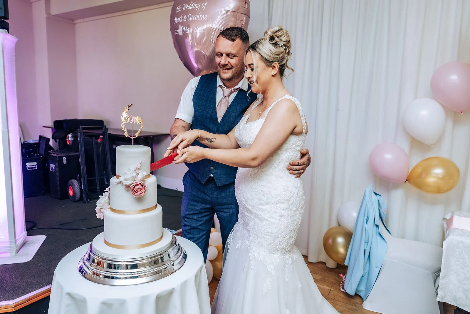 A smiling bride with a textured updo and a lace mermaid wedding dress cuts a three-tier white wedding cake alongside her groom in a blue tweed waistcoat, surrounded by pink and white balloons.