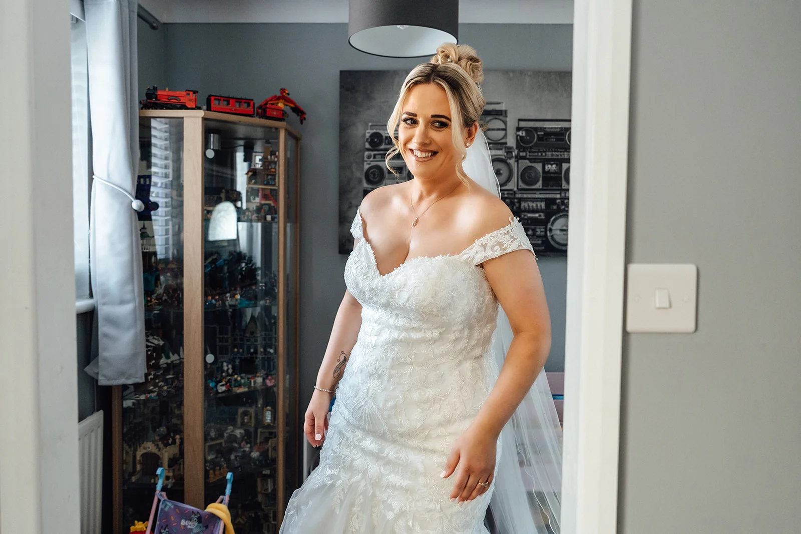 A smiling bride wearing a lace off-the-shoulder wedding gown and high bun stands in a bedroom, viewed through an open doorway.
