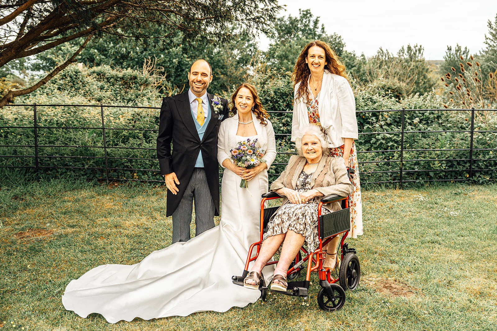 A newlywed couple poses outdoors in an English garden alongside family members, including a smiling grandmother seated in a red wheelchair.