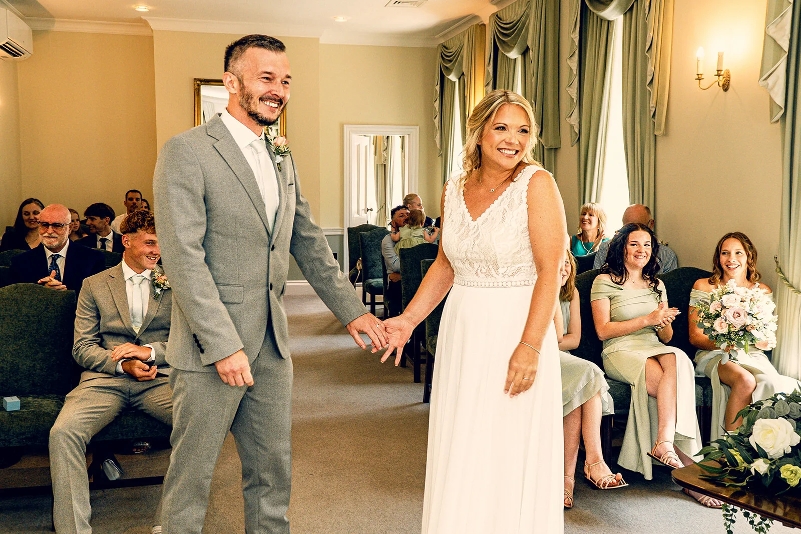 A smiling bride and groom holding hands and walking during their indoor wedding ceremony, surrounded by seated guests in an elegant hotel venue.