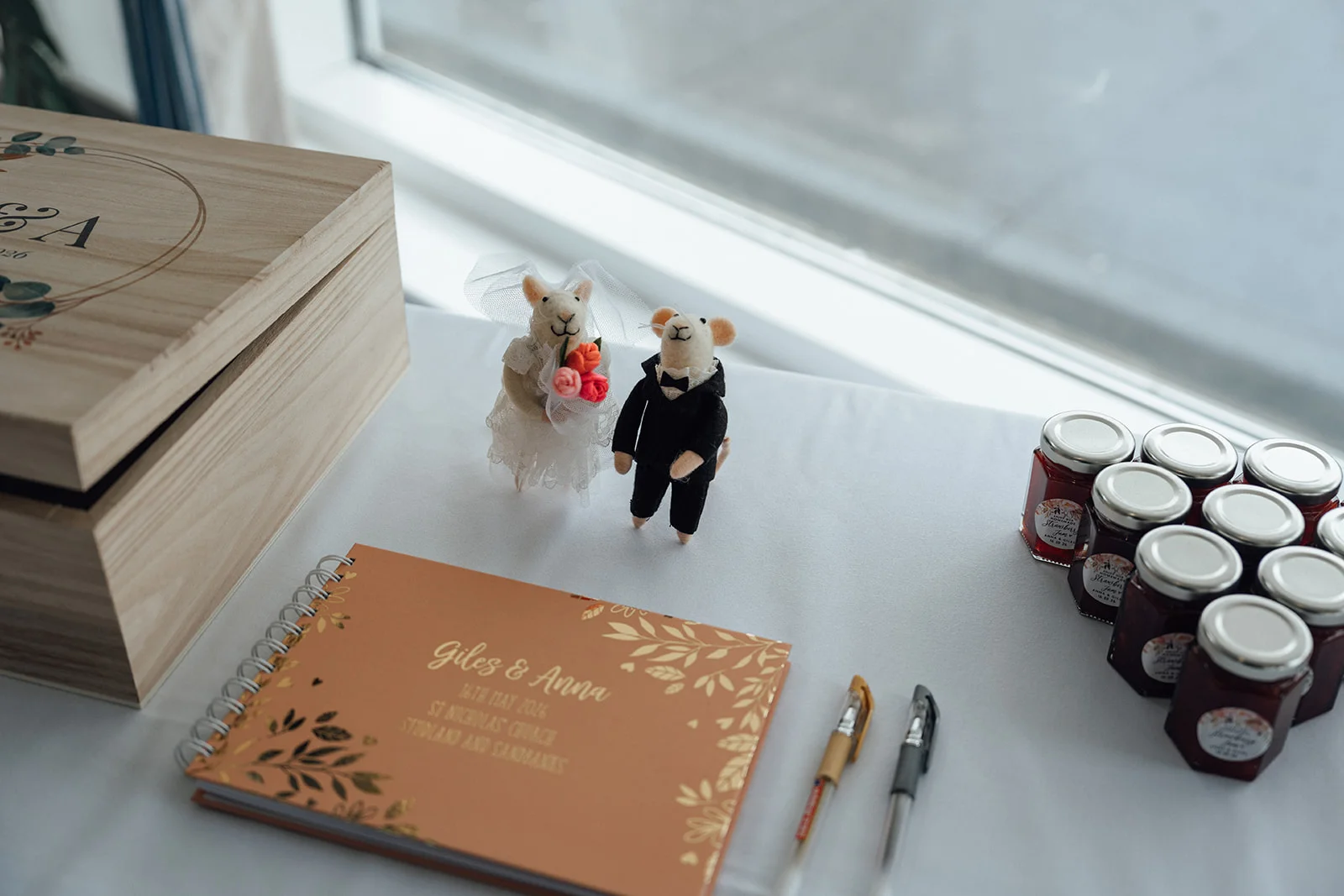 A rustic wedding guest book sits on a white table beside a wooden box, novelty mouse cake toppers, pens, and jars of jam favour gifts.