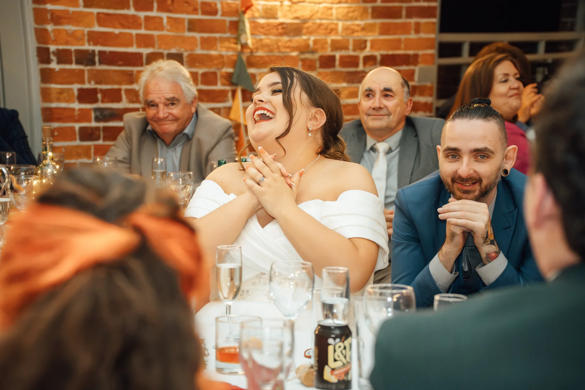 A joyful bride throws her head back in laughter during wedding speeches at a banquet table with exposed brick in the background.
