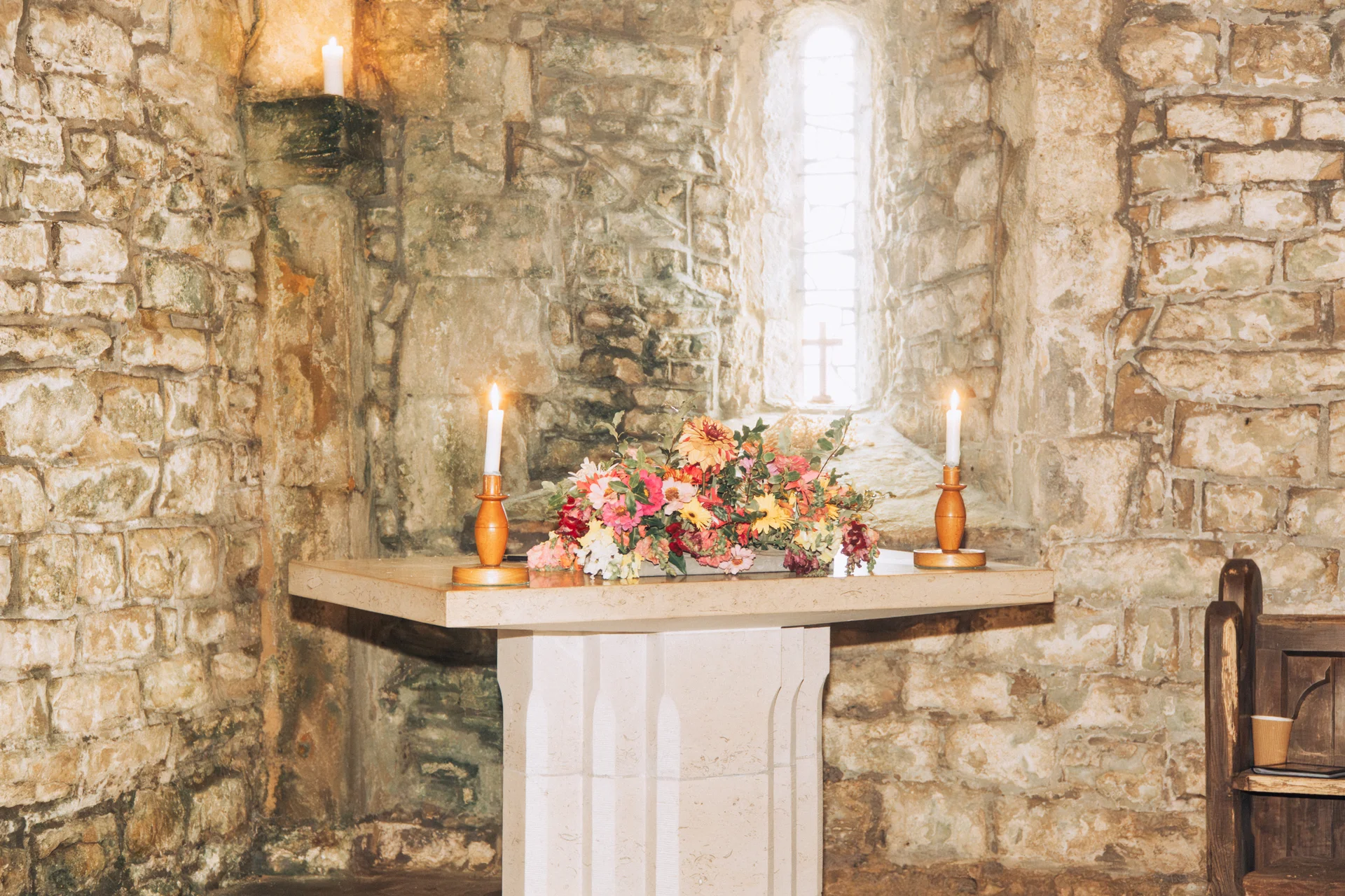 An altar in a historic stone church is adorned with colourful wildflowers and lit taper candles, with natural light streaming through a narrow stained glass window.