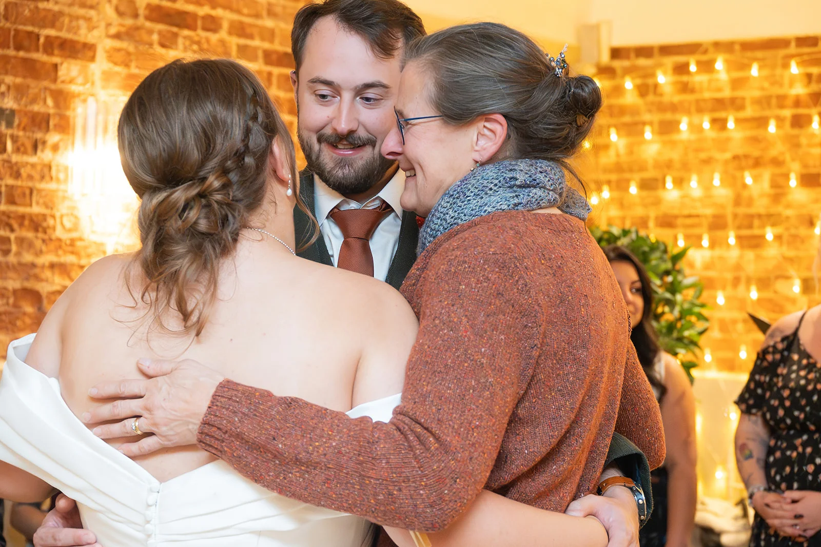An emotional guest embraces the newlywed couple in a warm, rustic venue featuring exposed brickwork and a glowing fairy light backdrop.